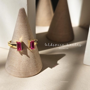 May include: Gold ring with two rectangular pink gemstones. The ring is on a wooden display stand. The text "Goldspoon Jewelry" and "Minimalist" is visible in the background.