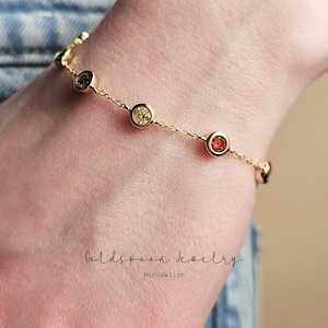 May include: A gold chain bracelet with small round gemstones in shades of yellow, orange, and red. The bracelet is on a person's wrist. The text "Goldspoon Jewelry Minimalist" is visible below the bracelet.