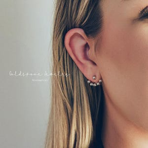 以下が含まれることがあります： 女性の耳のクローズアップ。2つのジュエリーが飾られています。1つ目は小さなスタッドピアスです。2つ目は、小さな丸い透明な石が連なったシルバーのイヤージャケットです。「Goldspoon Jewelry Minimalist」の文字が見えます。