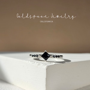 以下が含まれることがあります： 中央に黒い正方形の宝石、バンドの周りに小さな黒い宝石がついた銀の指輪。指輪は白い表面の上に置かれています。