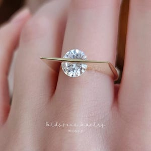 May include: A minimalist gold ring featuring a large, clear, oval-cut gemstone. The ring's design incorporates a flat, horizontal band that intersects the gemstone, creating a modern and unique aesthetic. The ring is labeled "Goldspoon Jewelry" and "Minimalist".