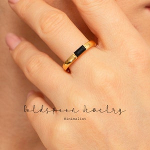 May include: A gold band ring with a black rectangular stone set in the center. The ring is worn on a person's finger. The text "Goldspoon Jewelry Minimalist" is visible below the ring.