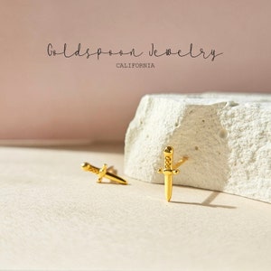 Pode incluir: Um par de brincos de adaga de ouro. Os brincos são pequenos e delicados, com um design simples. As adagas medem aproximadamente 1 cm de comprimento.