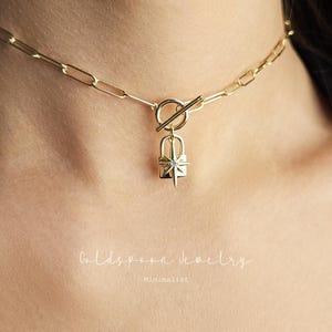 May include: A gold-colored necklace with a rectangular link chain and a toggle clasp. The pendant is a small, gold-colored padlock with a starburst design. The text "Goldspoon Jewelry" and "Minimalist" are visible.