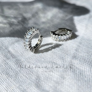May include: A pair of silver hoop earrings with a row of clear baguette-shaped cubic zirconia stones. The earrings are on a white fabric background. The text "Goldspoon Jewelry California" is visible below the earrings.