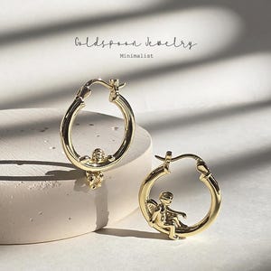 May include: A pair of gold hoop earrings featuring a small cherub figure attached to the bottom of each circular band. The text Goldspoon Jewelry Minimalist is visible at the top. The earrings have a polished metallic finish and a hinged clasp.