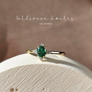 Puede incluir: Un anillo de oro con una piedra ovalada verde rodeada de diamantes más pequeños. El anillo está sobre una superficie blanca. Goldspoon Jewelry California.