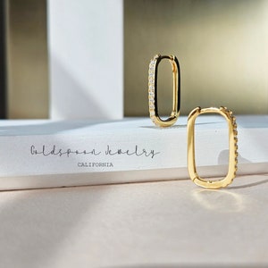 Puede incluir: Un par de pendientes de aro dorados con forma rectangular y una fila de pequeñas piedras transparentes alrededor del borde. Los pendientes están sobre una superficie blanca con el texto "Goldspoon Jewelry California" impreso en ella.