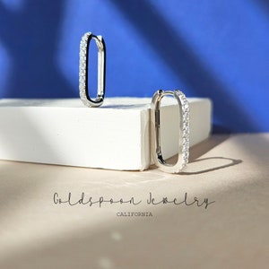 Puede incluir: Un par de pendientes de aro de plata con forma rectangular y una fila de piedras de circonita cúbica transparentes alrededor del borde. Los pendientes se muestran sobre una superficie blanca con un fondo azul.