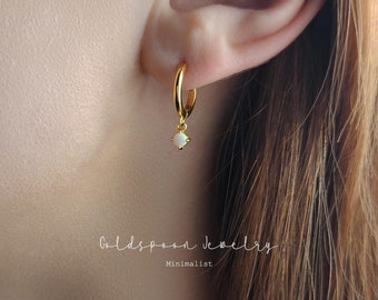 May include: Gold hoop earrings with a small white opal dangling from the bottom. The earrings are minimalist in style.