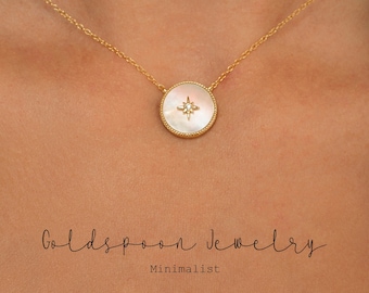 Puede incluir: Un collar de cadena de oro con un colgante redondo que presenta una incrustación de nácar blanco y una pequeña estrella de diamante en el centro. El colgante tiene un borde dorado con un borde texturizado. El collar está en el cuello de una modelo.