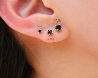 May include: Three black stud earrings of varying sizes (2mm, 3mm, and 4mm) are displayed on an ear. The earrings have a round, black gemstone set in a silver or white metal setting. The sizes are labeled next to each earring.