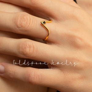 May include: A gold ring with a black gemstone in the shape of a V. The ring is on a person's finger. The text "Goldspoon Jewelry California" is visible below the ring.