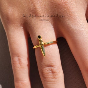 May include: Gold ring with a black stone and a dagger design. The ring is worn on a finger.