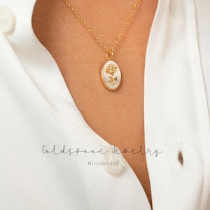 Könnte beinhalten: Eine goldene Kettenkette mit einem weißen ovalen Anhänger mit einem goldenen Blumendesign. Der Anhänger befindet sich an einem Modell, das ein weißes Hemd trägt. Der Text "Goldspoon Jewelry" und "Minimalist" ist unterhalb des Anhängers sichtbar.