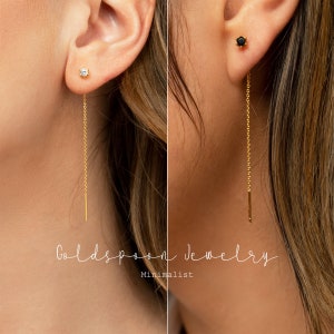 May include: Two close-up images of a person's ear wearing a gold chain earring with a small round gemstone at the top. The left image features a clear gemstone, while the right image features a black gemstone. The earrings are minimalist and delicate.
