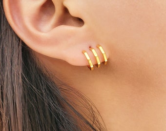 May include: Three small gold hoop earrings with a distinct hexagonal shape, worn in a row along the earlobe. Each earring features a polished metallic finish and a geometric design, creating a uniform and aligned appearance across the ear.