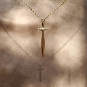 Könnte beinhalten: Eine goldene Halskette mit einem Schwertangänger. Das Schwert ist mit kleinen, klaren Steinen verziert.