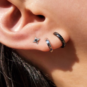 May include: A person's ear with three earrings. The top earring is a small silver star with a black stone in the center. The bottom two earrings are silver hoops, one is black and the other is silver.