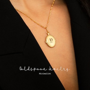 Puede incluir: Un collar de cadena de oro con un colgante de medallón ovalado de oro con una letra "S" grabada en negro. El medallón está en un modelo que lleva una chaqueta negra. El texto "Goldspoon Jewelry" y "Minimalist" es visible debajo del modelo.