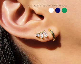 May include: A close-up of a person's ear wearing a gold hoop earring and a gold stud earring with a row of baguette-shaped clear stones. The text "AVAILABLE IN WHITE, EMERALD & SAPPHIRE CZ" is above the image. Three color swatches are shown: white, dark blue, and green.