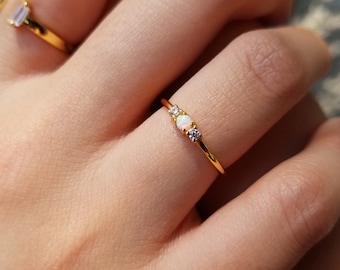 May include: A gold ring with three gemstones: a central opal and two smaller clear stones. The ring is on a finger, and another gold ring is visible in the background. The ring's band is thin and delicate.