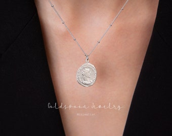 May include: A silver chain necklace with a silver coin pendant featuring a profile of a woman. The necklace is on a person's chest, and the background is black.