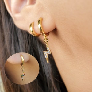 May include: Gold hoop earrings with a dangling lightning bolt charm. The charm is set with clear cubic zirconia stones.