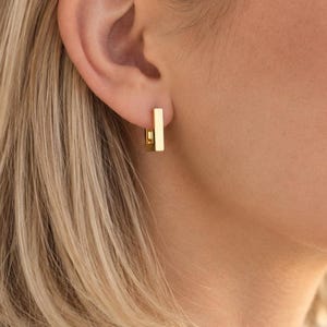 May include: A small gold hoop earring featuring a vertical rectangular bar charm. The earring is worn in the earlobe and has a polished metallic finish. The design consists of a thin circular hoop connected to a straight, flat, rectangular gold bar.