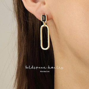 May include: Gold-toned earrings with a rectangular black stone at the top and a long, open oval shape hanging below. The earrings are minimalist in design. The text "Goldspoon Jewelry" and "Minimalist" are visible.