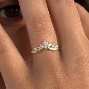 May include: A gold V-shaped ring featuring a central round brilliant-cut stone flanked by smaller marquise-cut stones arranged in a crown-like pattern along the band. The metal band has a polished finish and follows a chevron contour design.