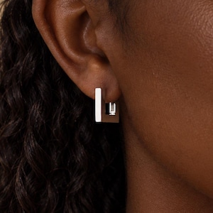May include: A small, polished silver-tone hoop earring with a distinct square shape. The earring features a flat, rectangular profile that forms a geometric loop, securely fastened through the earlobe with a visible hinge mechanism at the base.