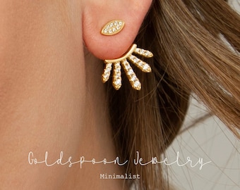 May include: Gold earrings with a sunburst design and a small oval-shaped earring with clear stones. The earrings are minimalist and delicate.