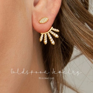 May include: Gold earrings with a sunburst design and a small oval-shaped earring with clear stones. The earrings are minimalist and delicate.