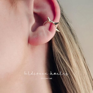 Könnte beinhalten: Ein goldfarbener Ohrklemme mit Stern-Design. Der Ohrschmuck wird am Ohr getragen und weist mehrere sternförmige Akzente auf. Der Schmuck ist minimalistisch und elegant, perfekt, um einen Hauch von Glanz zu verleihen.