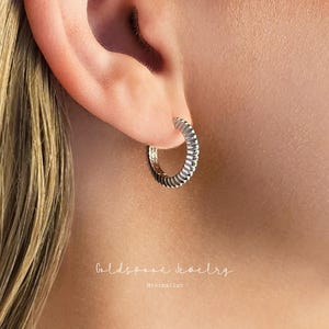 Puede incluir: Primer plano de un pendiente de aro plateado con un diseño acanalado. El pendiente está en la oreja de una persona. La imagen también incluye el texto "Goldspoon Jewelry" y "Minimalist".