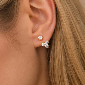 May include: Two gold stud earrings worn in the earlobe. One earring features a single round clear stone, while the other is a cluster of three round clear stones arranged in a triangular shape. Both earrings have a polished gold metal setting.