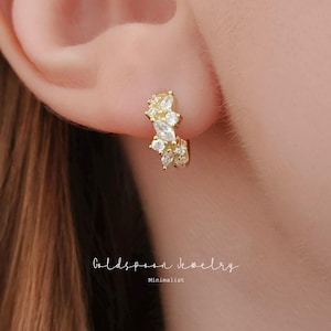 May include: A gold-colored hoop earring with a cluster of clear and yellow gemstones. The earring is worn on an ear. The text "Goldspoon jewelry" and "Minimalist" are visible in the image.