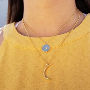May include: Two gold necklaces with pendants. The top necklace has a round pendant with a blue background and a gold star with white diamonds. The bottom necklace has a crescent moon pendant with white diamonds.