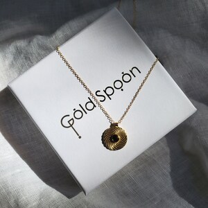 Op de afbeelding: Een gouden ketting met een zwarte en gouden swirl hanger. De ketting wordt gepresenteerd op een witte doos met de tekst "Goldspoon" erop gedrukt.