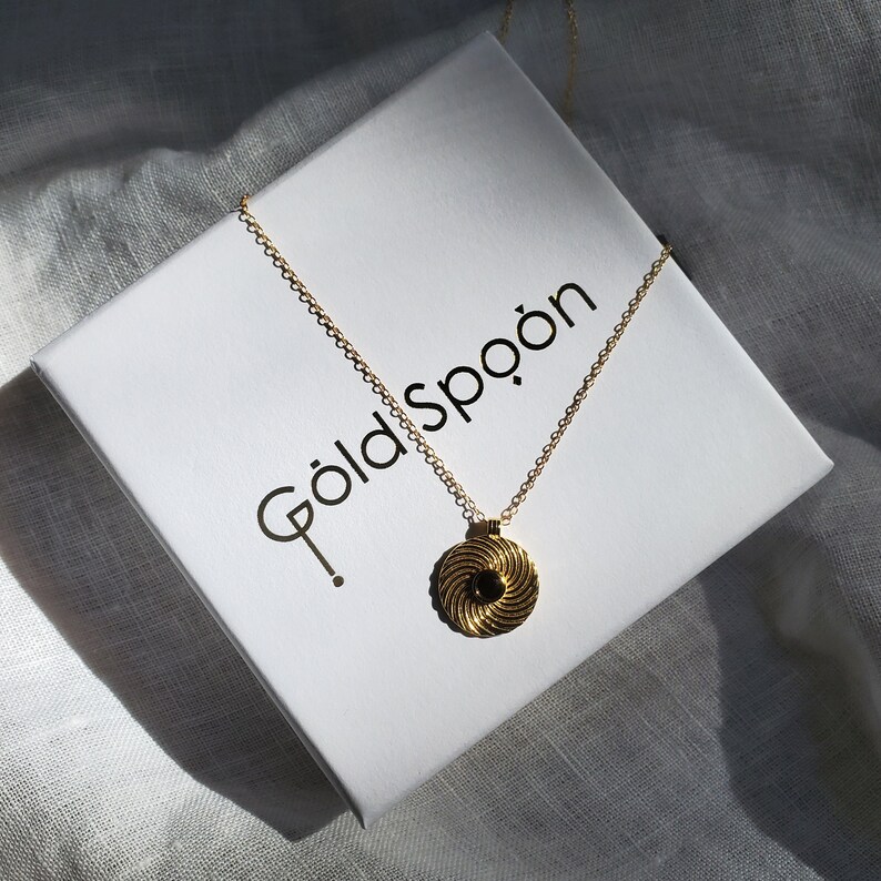 Op de afbeelding: Een gouden ketting met een zwarte en gouden swirl hanger. De ketting wordt gepresenteerd op een witte doos met de tekst "Goldspoon" erop gedrukt.