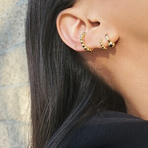 May include: A close-up of a person's ear with three gold hoop earrings. The earrings are all different sizes and styles. The largest earring is a beaded hoop, the middle earring is a simple hoop, and the smallest earring is a stud.