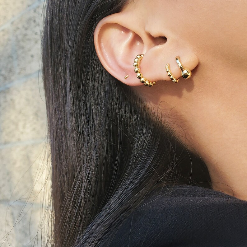 May include: A close-up of a person's ear with three gold hoop earrings. The earrings are all different sizes and styles. The largest earring is a beaded hoop, the middle earring is a simple hoop, and the smallest earring is a stud.