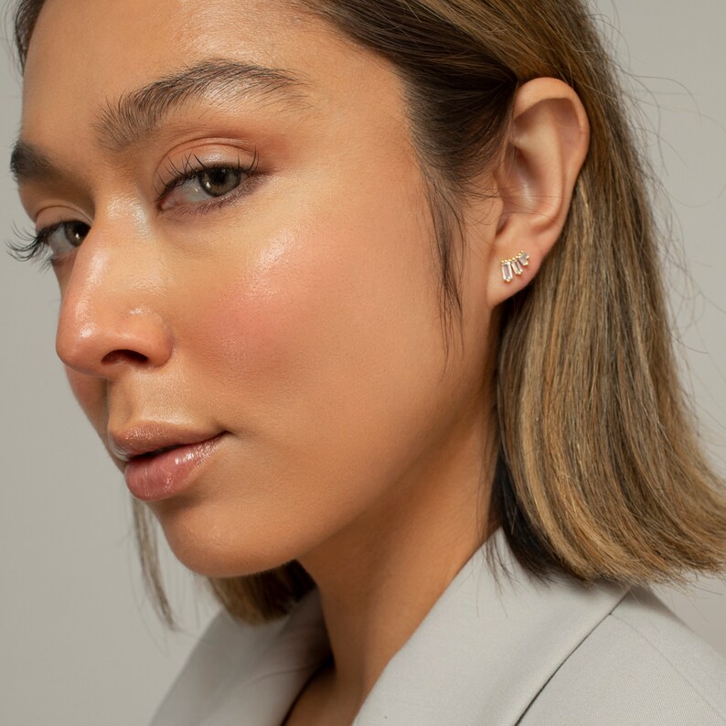 May include: A close-up of a person's ear wearing a gold earring with small, clear baguette-shaped stones. The earring is a cluster of stones set in a curved shape.