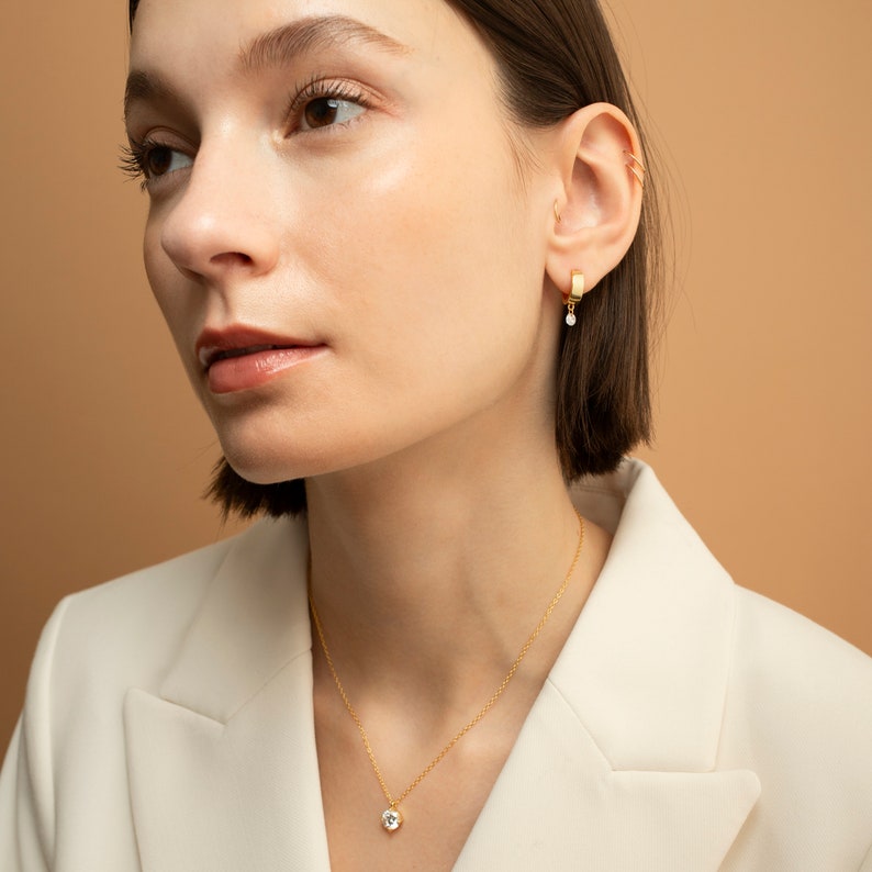 May include: A woman wearing a white blazer with a gold chain necklace featuring a small round diamond pendant. She is also wearing a gold hoop earring with a small diamond dangling from it.