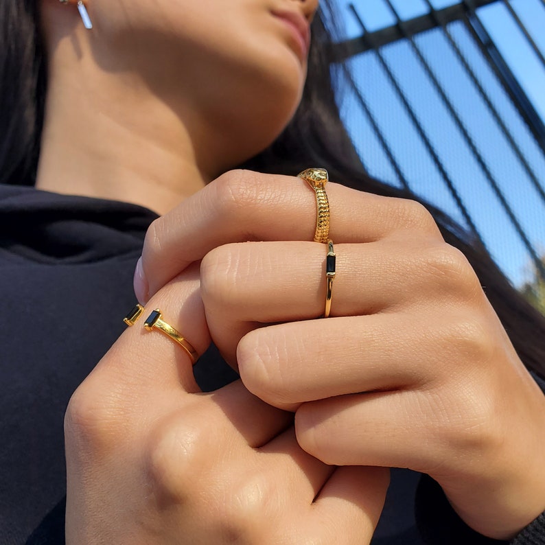 May include: A hand wearing three gold rings with black gemstones. The rings are stacked on the middle finger.