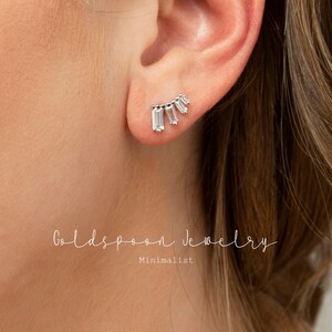 May include: A pair of silver earrings with a row of five clear baguette-shaped stones. The earrings are in the shape of a crescent moon. The earrings are minimalist and delicate.