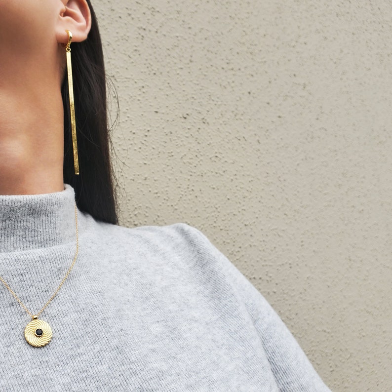 Op de afbeelding: Een gouden ketting met een zwarte ronde hanger met een swirl-ontwerp. De ketting wordt gedragen door een persoon met lang donker haar en een grijze trui met een hoge hals. De persoon draagt een lange gouden staaf oorbel.