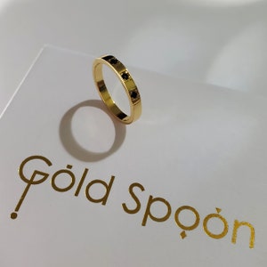 Op de afbeelding: Een gouden ring met vier zwarte vierkante stenen die in de band zijn gezet. De ring ligt op een wit oppervlak met de tekst "Gold Spoon" in gouden letters.
