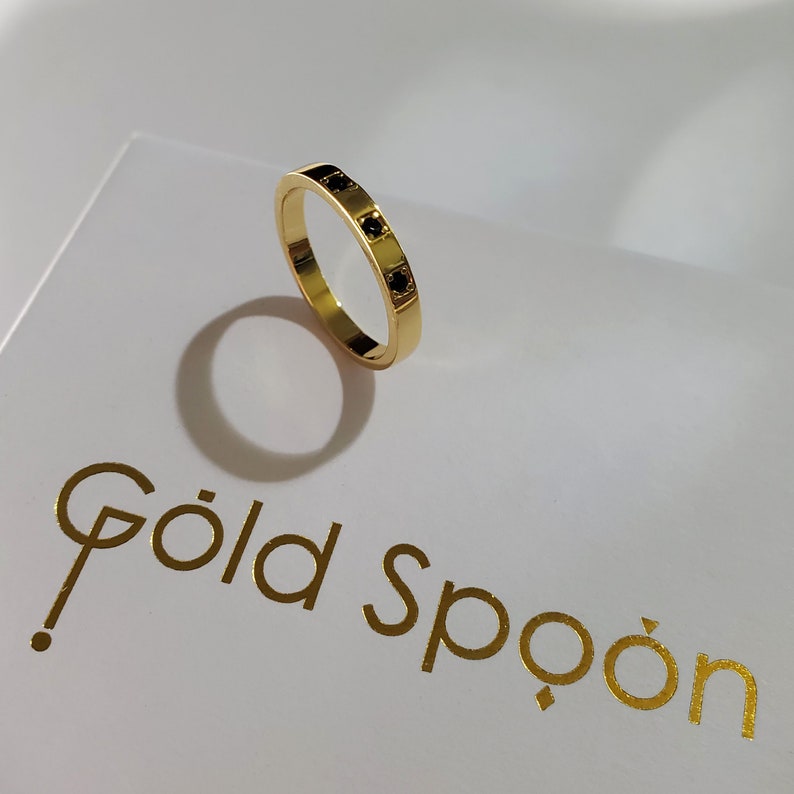 Op de afbeelding: Een gouden ring met vier zwarte vierkante stenen die in de band zijn gezet. De ring ligt op een wit oppervlak met de tekst "Gold Spoon" in gouden letters.